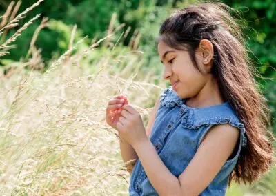 Natural portrait of a young girl with long brown hair, wearing a denim dress, gently examines a blade of tall grass in a sunny outdoor setting.