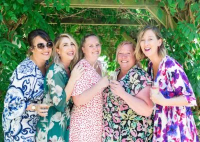 Five smiling women holding champagne glasses and celebrating outdoors, standing under a leafy green pergola on a sunny day. They are dressed in colorful, patterned summer dresses.