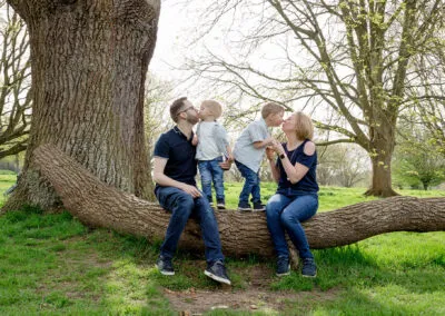 A playful family portrait captured outdoors, with parents and their two young children sitting on a fallen tree, exchanging kisses.