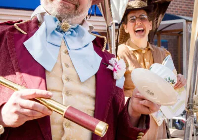 A vibrant portrait captures the playful spirit of a costumed character, beaming with infectious enthusiasm. Adorned in period attire, complete with a dashing top hat and telescope, he perfectly embodies the joy of storytelling and imaginative play. In the background, a companion's genuine laughter adds to the scene's delightful charm.