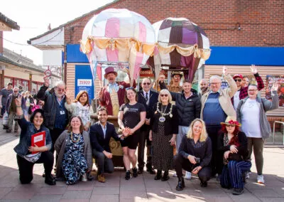 This lively group portrait captures the vibrant energy of a community event. Attendees, some dressed in character, are all smiling and waving around two impressive hot air balloon displays, set against a backdrop of local shops.