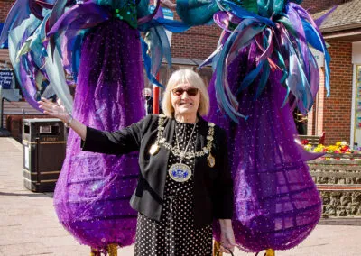 This lively photograph captures an engaging moment between the mayor of Rainham and two captivating stilt walkers. The performers' elaborate, colourful attire and impressive height draw the eye, while the woman's warm expression suggests a delightful interaction at an outdoor event.