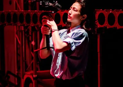 Behind the scenes capturing the intensity of the moment, a filmmaker focuses on their craft amidst a striking red-lit stage. The subtle glow illuminates both the artist and the intricate audio setup, hinting at the dynamic energy of a live performance.